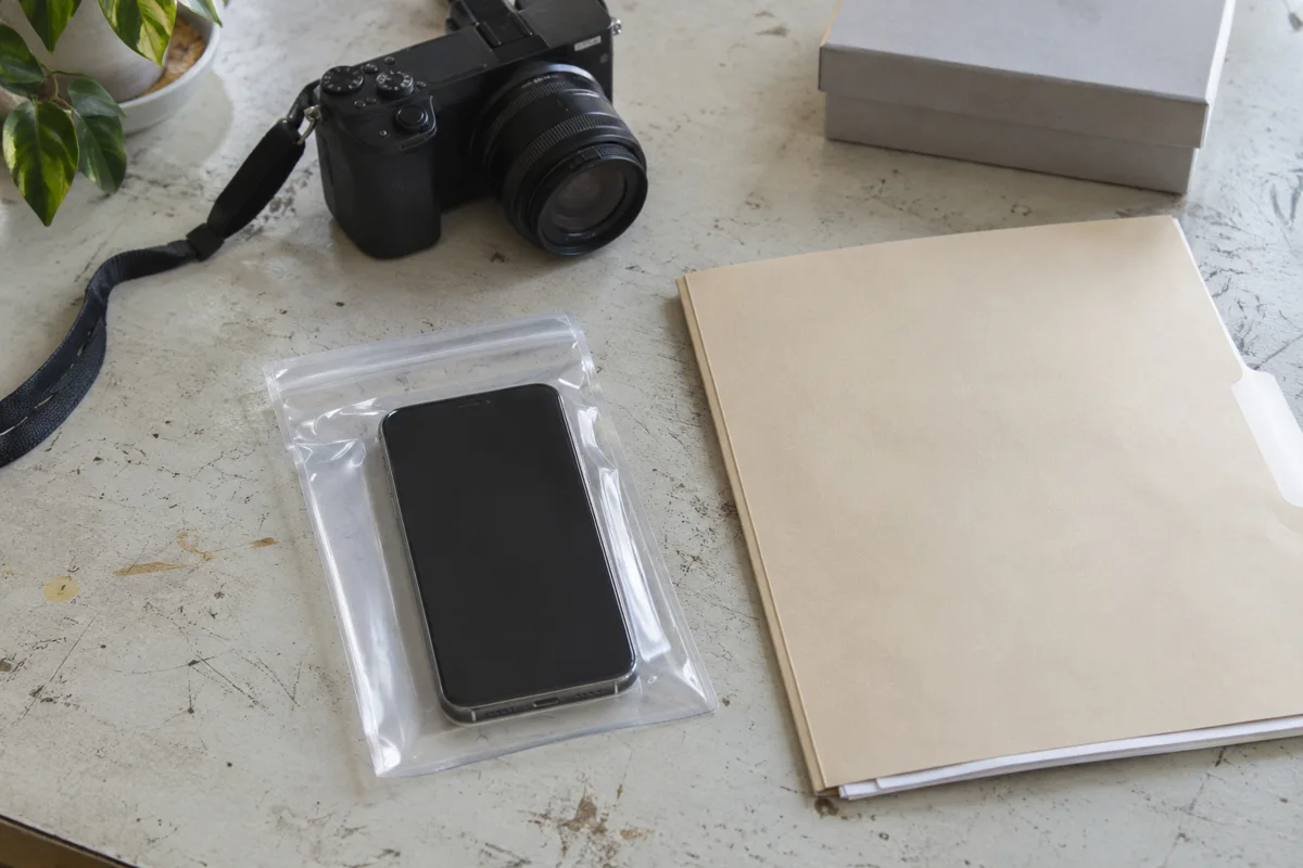 A dark-screen phone in a clear pouch beside a blank folder, camera and closed box.